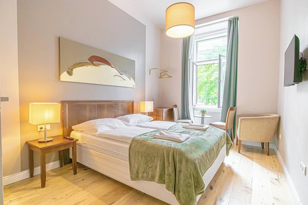 A neatly arranged bedroom with a bed, bedside lamps, a chair, a window with curtains, and a wall-mounted TV.