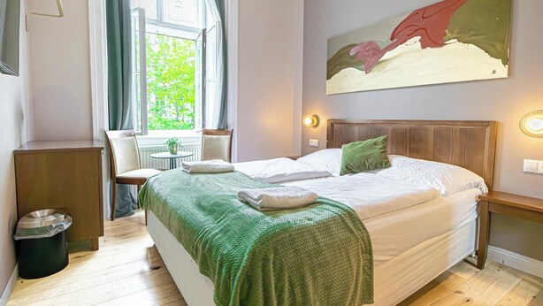 A hotel room with a large bed, a green blanket, a wooden headboard, a painting on the wall, and a window with curtains. - Boutique hotel (Vienna)
