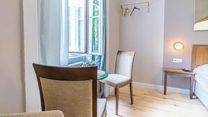 A room with a wooden floor, a wicker chair, a wooden table, a bed, a window with curtains, and a wall-mounted light fixture. - Boutique hotel (Vienna)