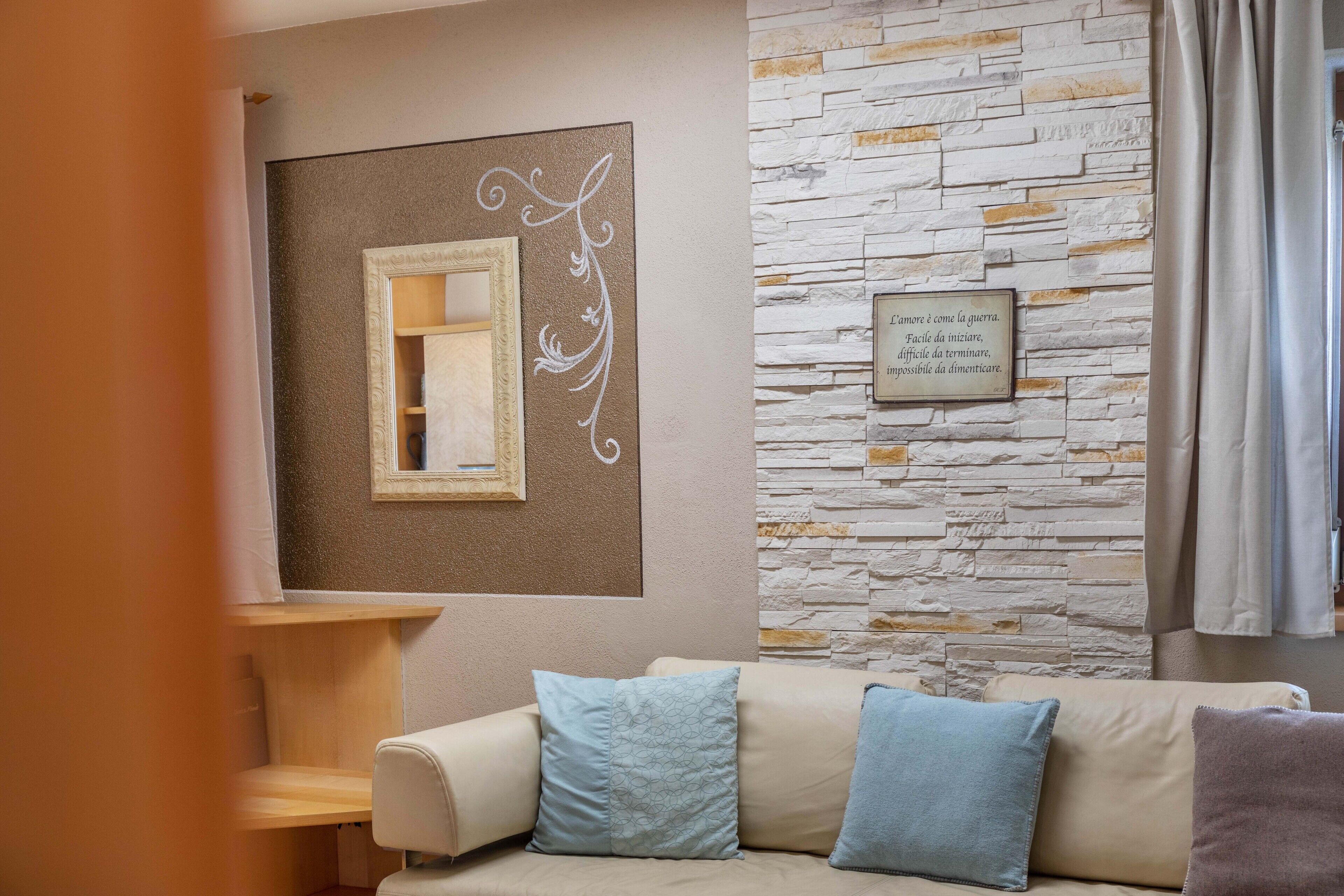 A living room with a beige sofa, patterned cushions, a mirror, and a stone wall with a framed quote.