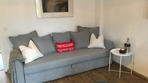 Un canapé gris agrémenté de plusieurs coussins, une petite table ronde avec une bouteille et des verres, et un tableau encadré accroché au mur.
