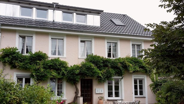 Un bâtiment à deux étages, doté d’un toit plat, de nombreuses fenêtres et d’un jardin agrémenté de fleurs et d’arbustes.