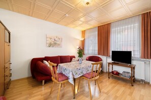 Un salon chaleureux avec un plancher en bois, un canapé rouge, une table à manger avec des chaises, un téléviseur posé sur un meuble et une fenêtre avec des rideaux.