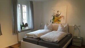 A bedroom with a bed, a window with curtains, a radiator, a wall-mounted lamp, and a painting. - Apartment 'Unterm Blauregen' with Private Terrace and Wi-Fi (Öhningen)