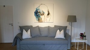 A living room with a grey sofa, a wall-mounted abstract painting, a side table with a lamp, and a door. - Apartment 'Unterm Blauregen' with Private Terrace and Wi-Fi (Öhningen)