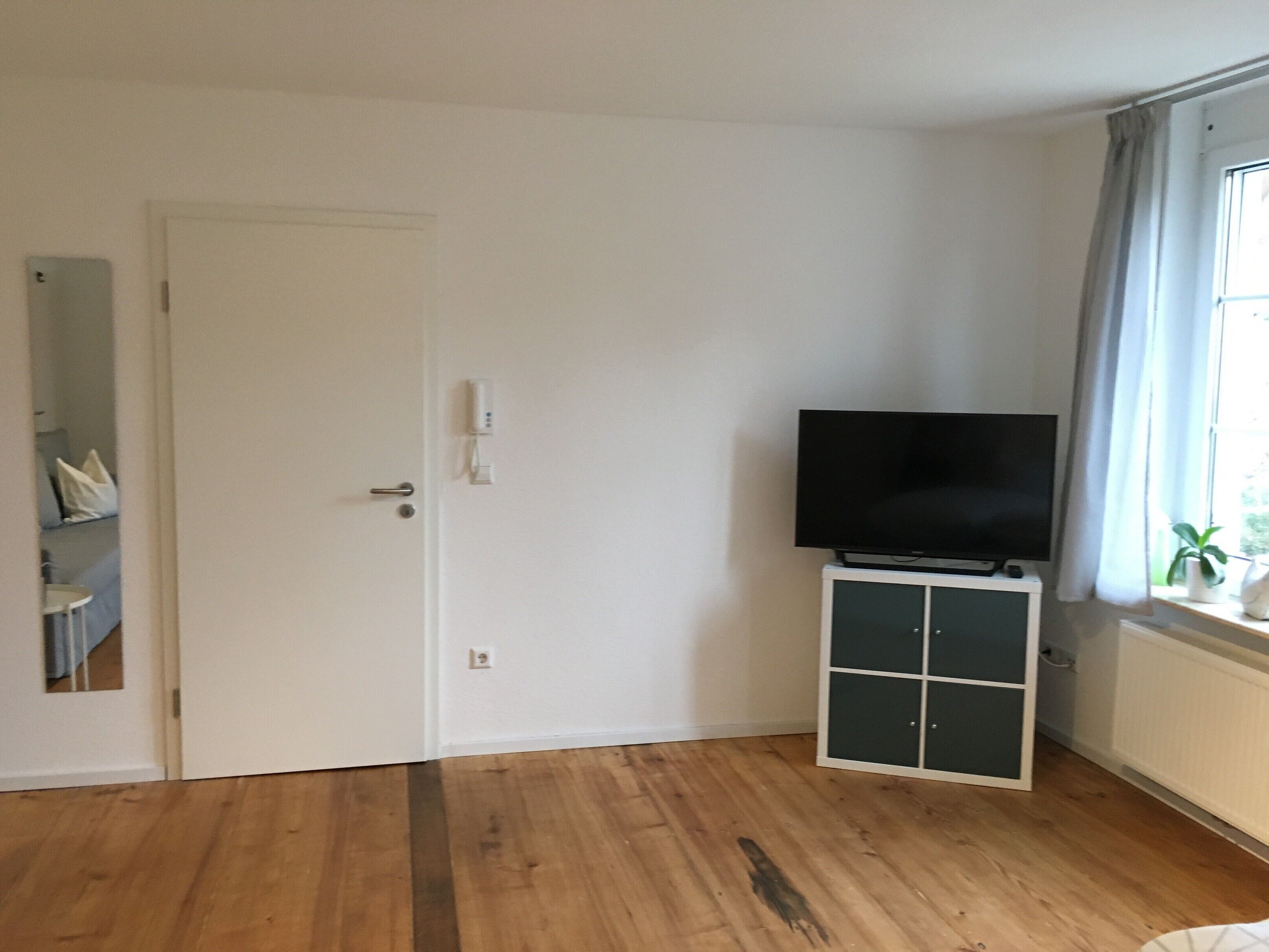 Ein Zimmer mit Holzboden, einer weißen Tür, einem Fernseher auf einem Schrank und einem Fenster mit Vorhängen.