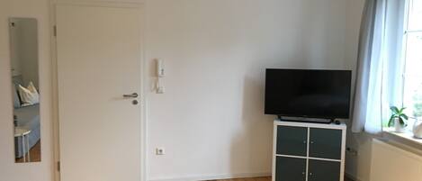 Ein Zimmer mit Holzboden, einer weißen Tür, einem Fernseher auf einem Schrank und einem Fenster mit Vorhängen.