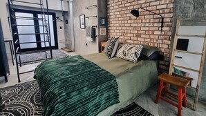 Een slaapkamer met een bakstenen muur, een bed met een groene deken, een houten ladder en een klein krukje.