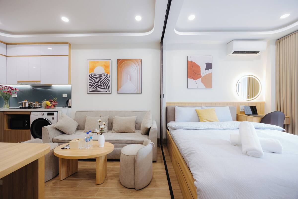 Una habitación de hotel moderna con sofá, una mesa redonda de madera, una cama con cabecera, un escritorio con una laptop y una lavadora en la esquina.