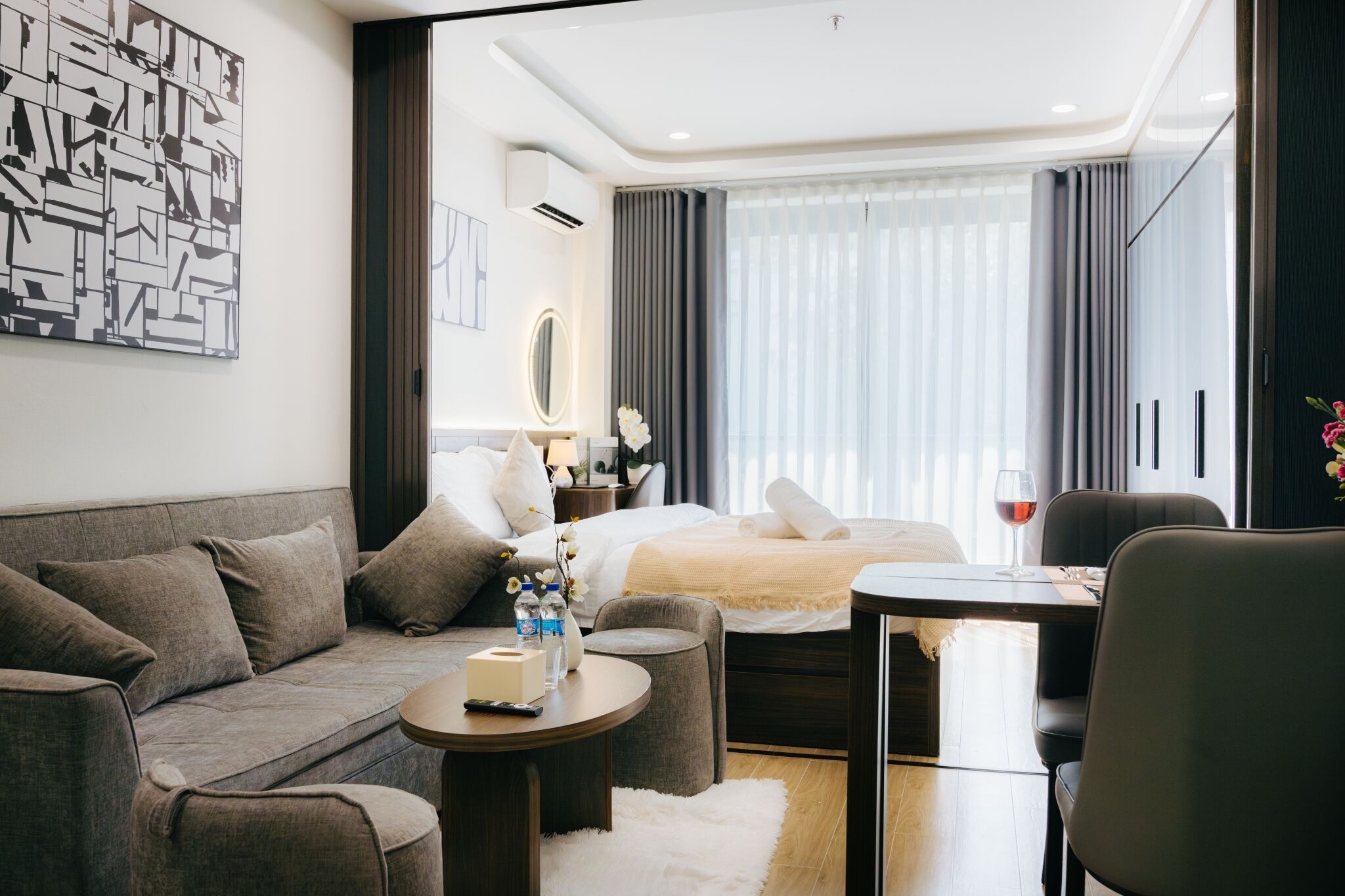 A modern hotel room with a sofa, a small round table, a bed with a white duvet, a nightstand with a lamp, and a view of the city through large windows.