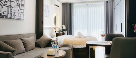 A modern hotel room with a sofa, a small round table, a bed with a white duvet, a nightstand with a lamp, and a view of the city through large windows.