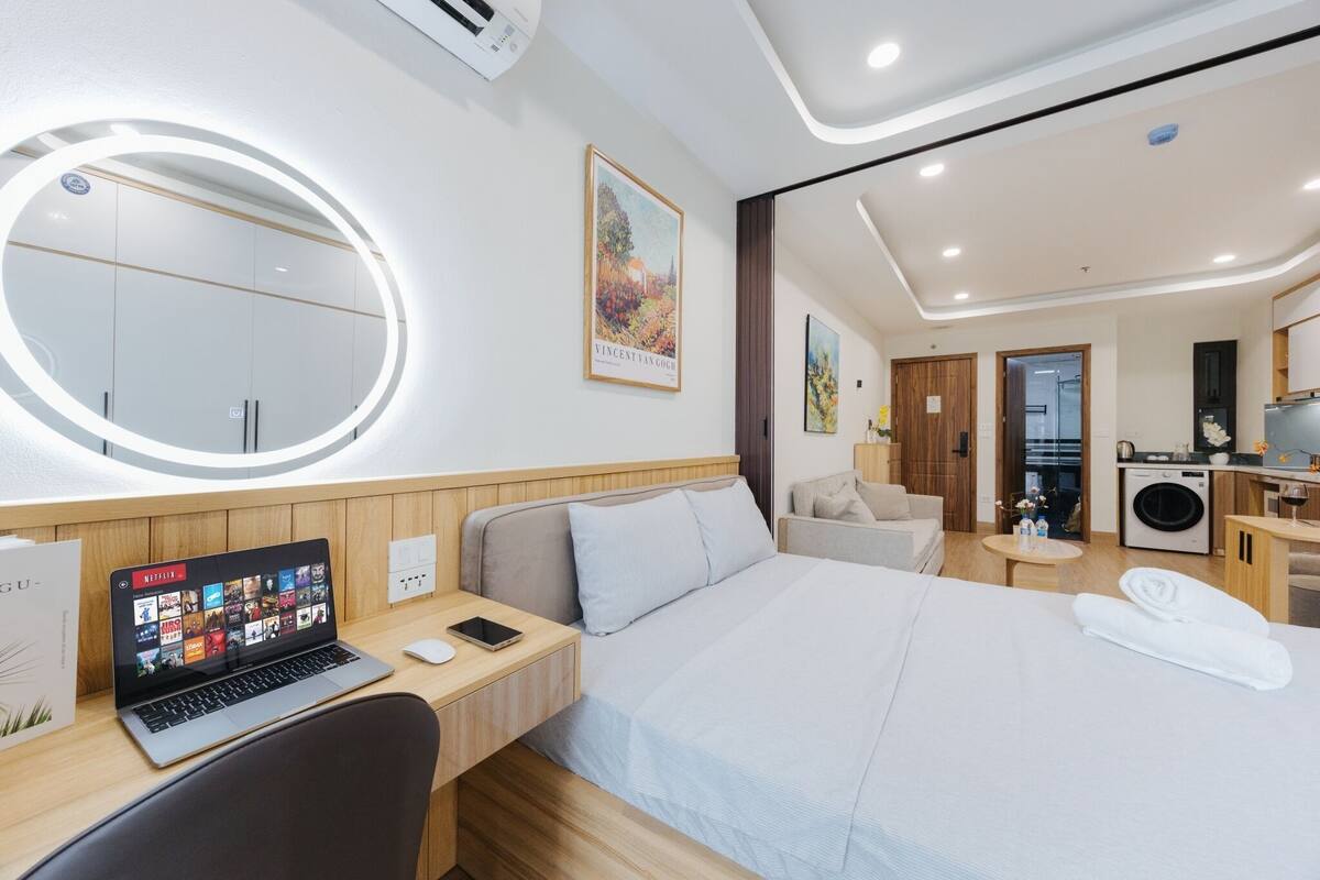 Una habitación de hotel moderna con cama, escritorio con una computadora portátil, espejo redondo, pequeño espacio para sentarse y kitchenette.