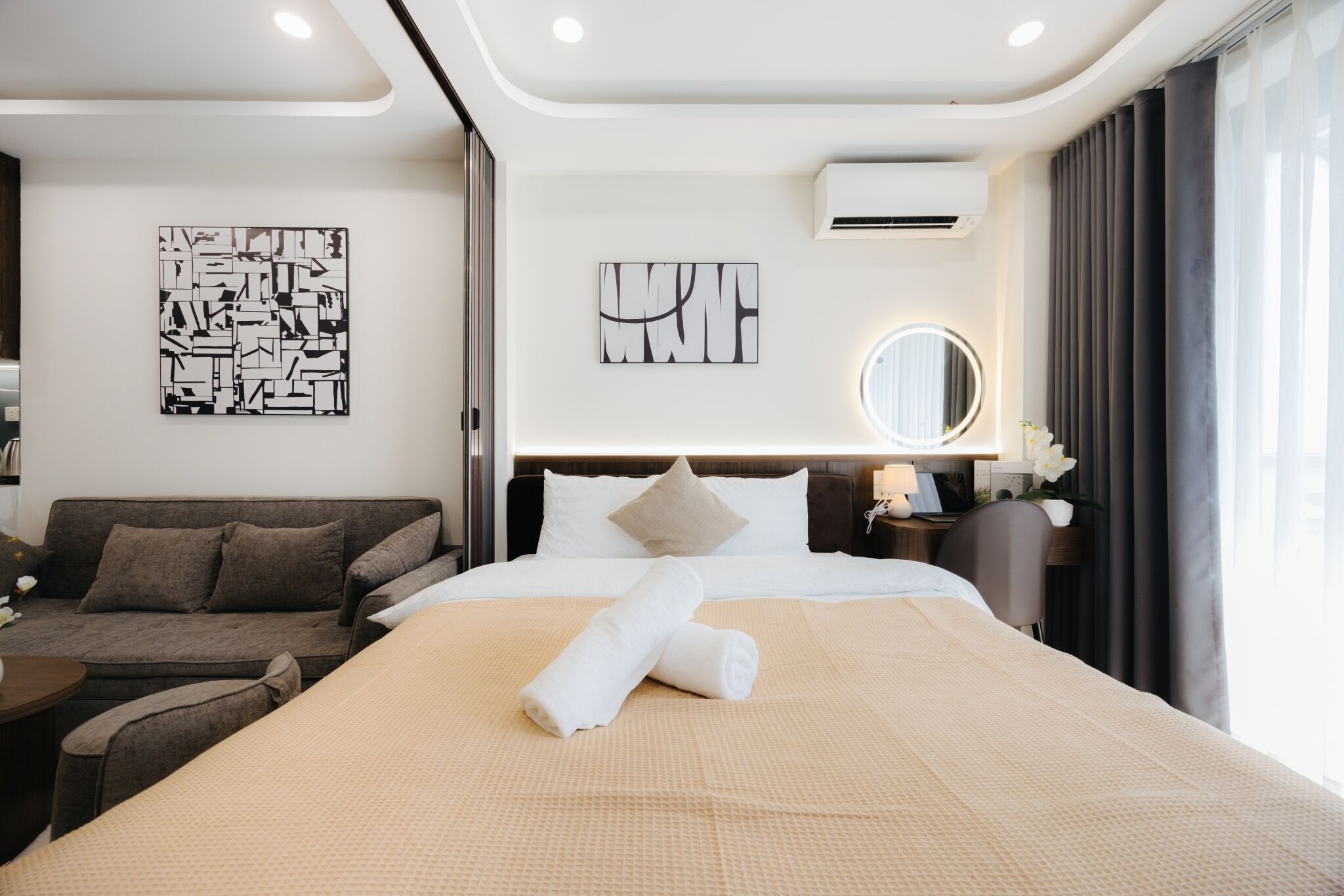 A modern hotel room with a large bed, a sofa, a round mirror, and artwork on the walls.