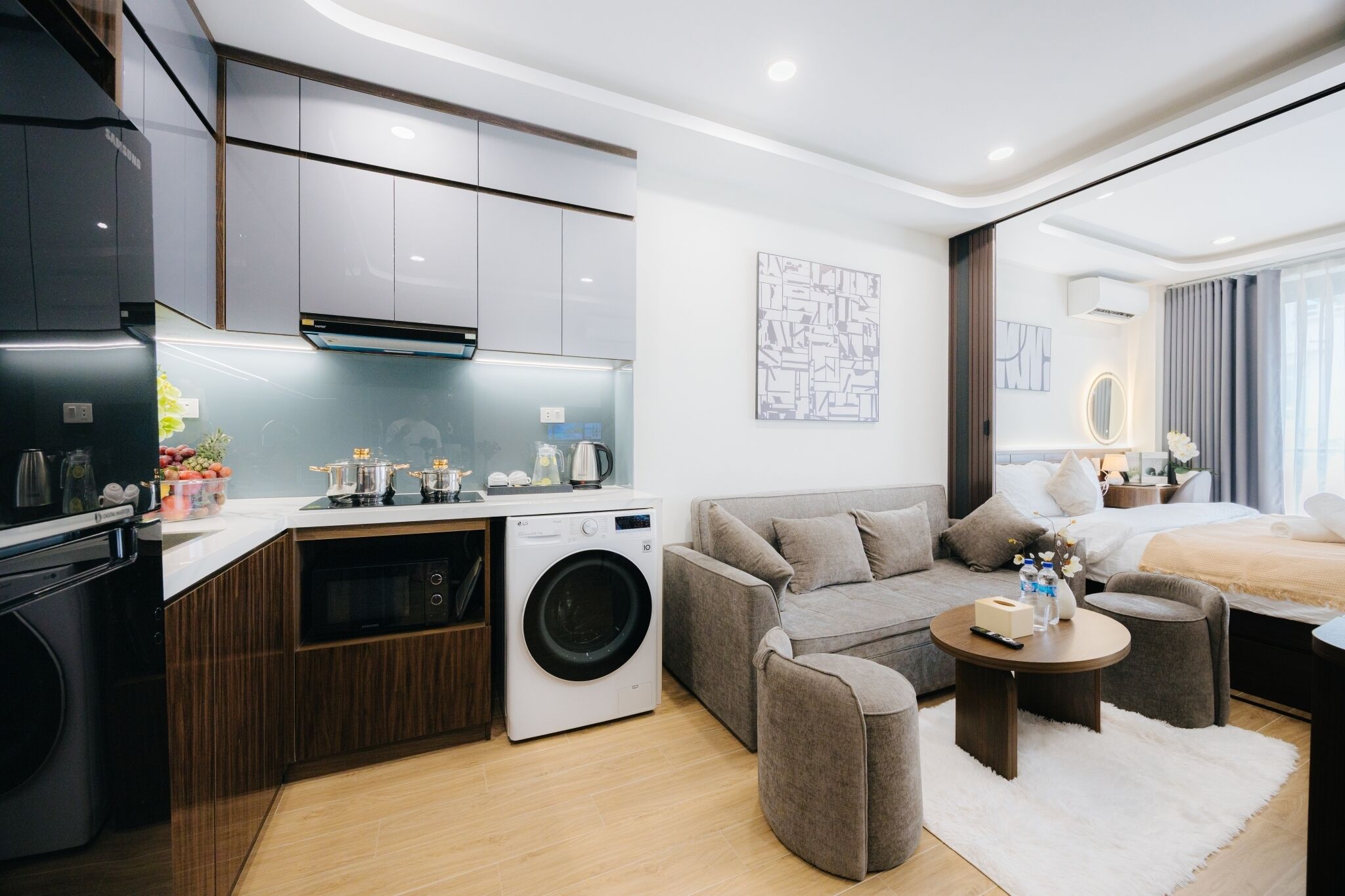 A modern hotel room with a kitchenette, a washing machine, a sofa, a coffee table, and a bed.