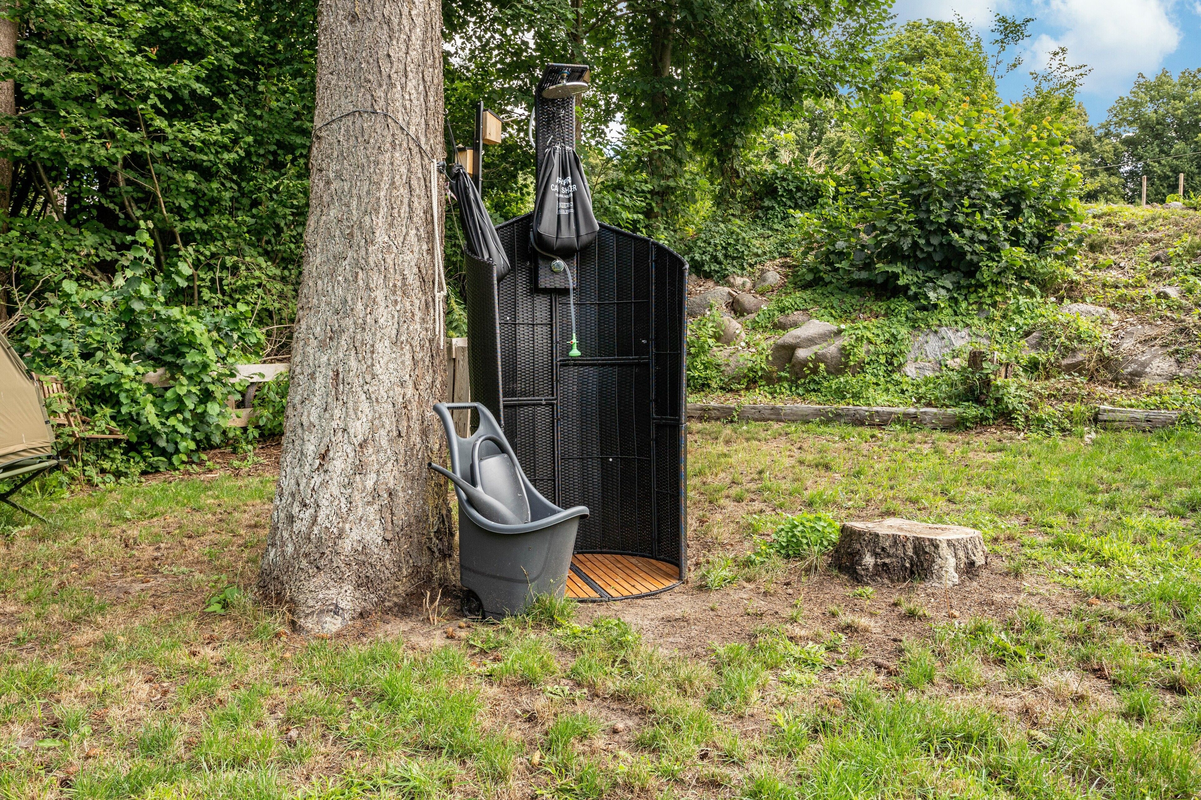 Une douche extérieure noire, constituée d’une structure et dotée d’un siège en bois et d’un sac suspendu, le tout entouré de verdure et d’un arbre.