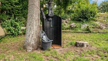 Une douche extérieure noire, constituée d’une structure et dotée d’un siège en bois et d’un sac suspendu, le tout entouré de verdure et d’un arbre.