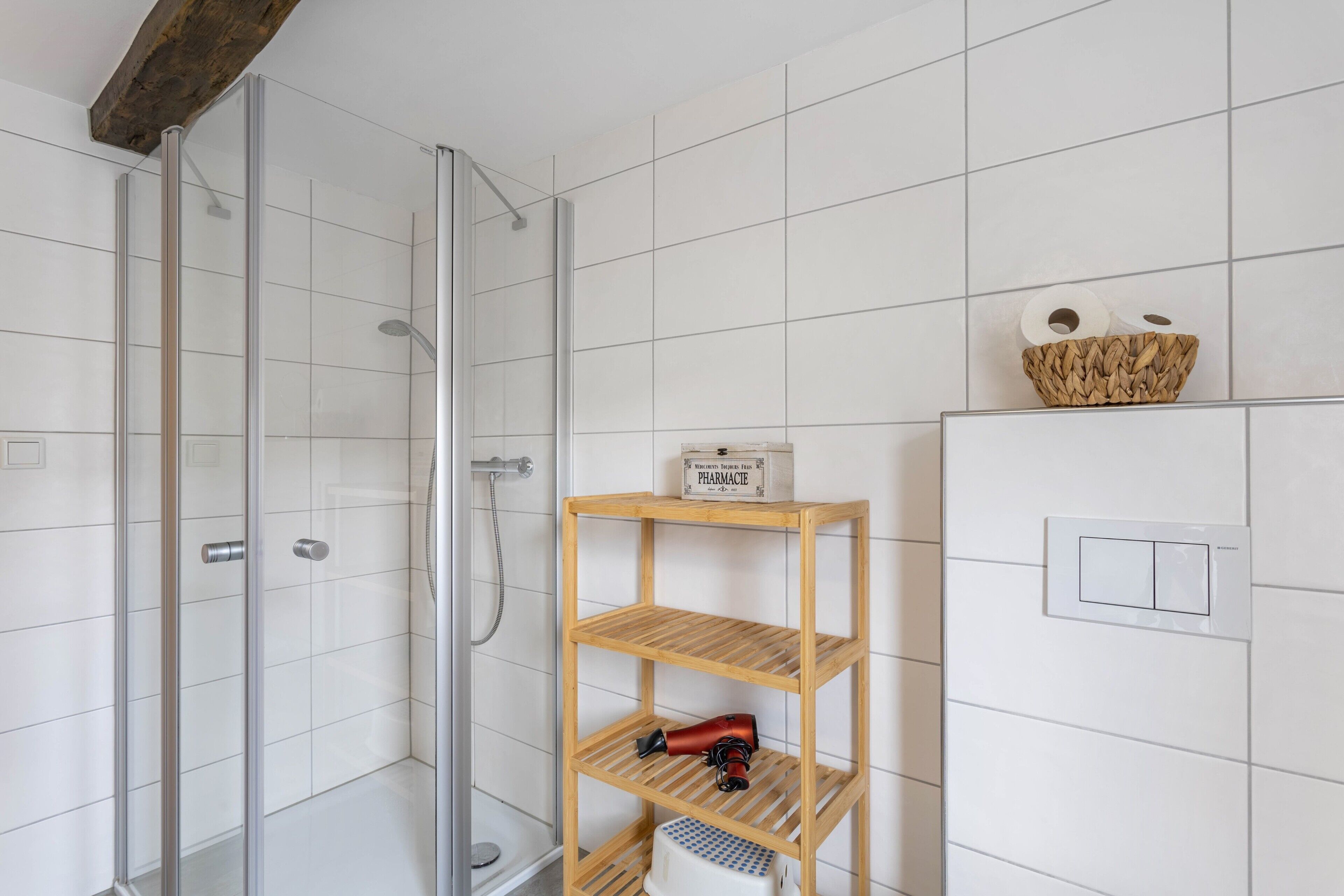 Een douche met glazen deuren, een houten plank met een doosje met de tekst 'PHARMACIE' en een mand aan de muur.