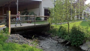 Um edifício com área externa coberta, uma ponte sobre um riacho, e uma pessoa caminhando.