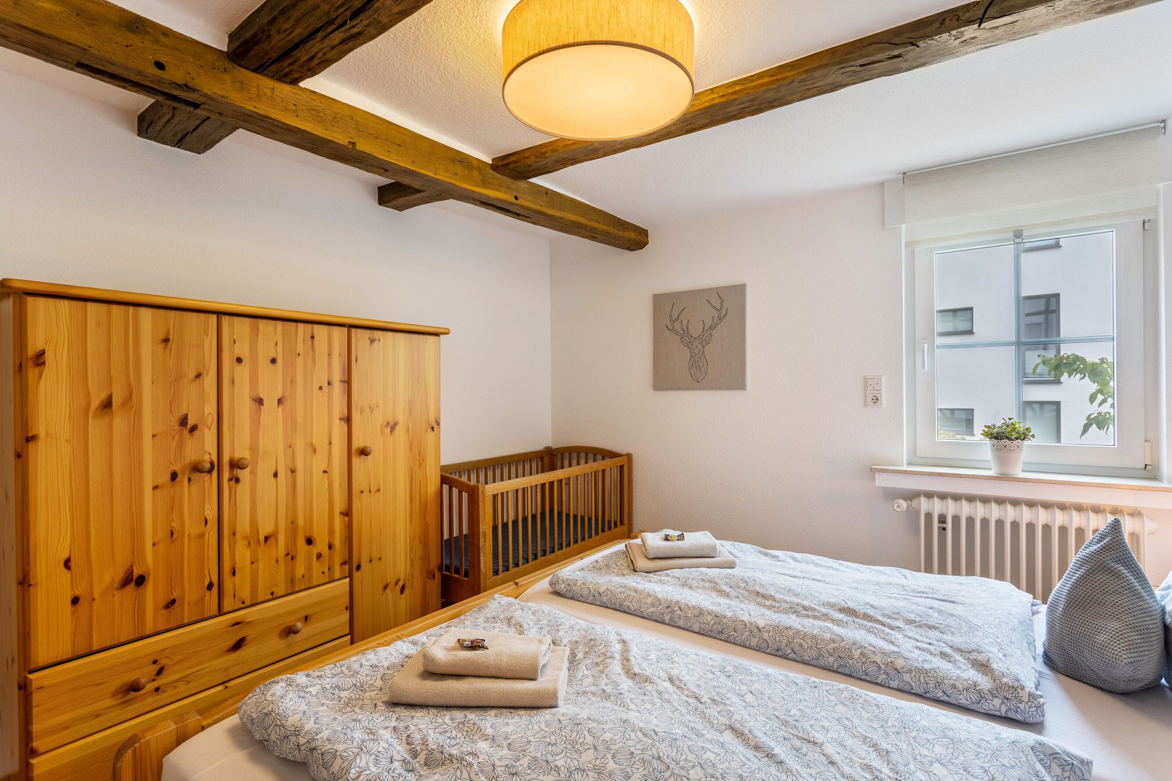 Ein Schlafzimmer mit zwei Betten, einem Holzschrank, einem Babybett, einem Heizkörper und einem Fenster mit einer Pflanze.