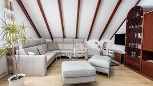A modern living room with a sectional sofa, a glass coffee table, a white ottoman, a wooden cabinet with glassware, and a potted plant. - Holiday Apartment 'Waldmann' with Shared Terrace, Garden, and Wi-Fi (Hattorf am Harz)