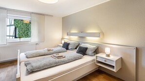 A bedroom with a bed, bedside tables, a lamp, a window with a view of greenery, and a ceiling light.