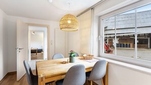 Un comedor con una mesa de madera, sillas grises, una lámpara colgante de fibra y vistas a la calle a través de grandes ventanales.