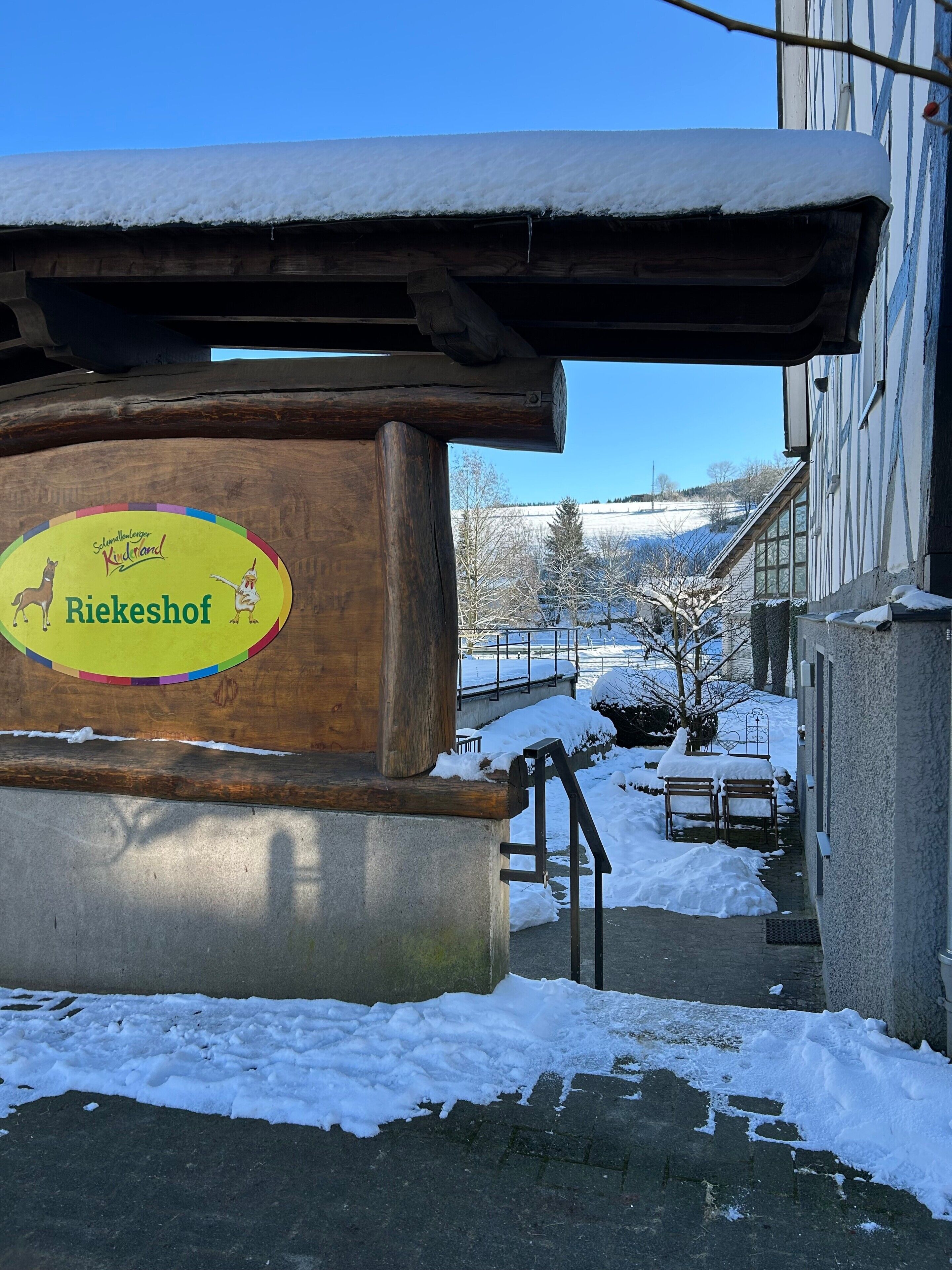 Un panneau en bois portant l’inscription « Riekeshof » et une illustration de cheval, un banc recouvert de neige et un bâtiment doté d’un toit en pente.