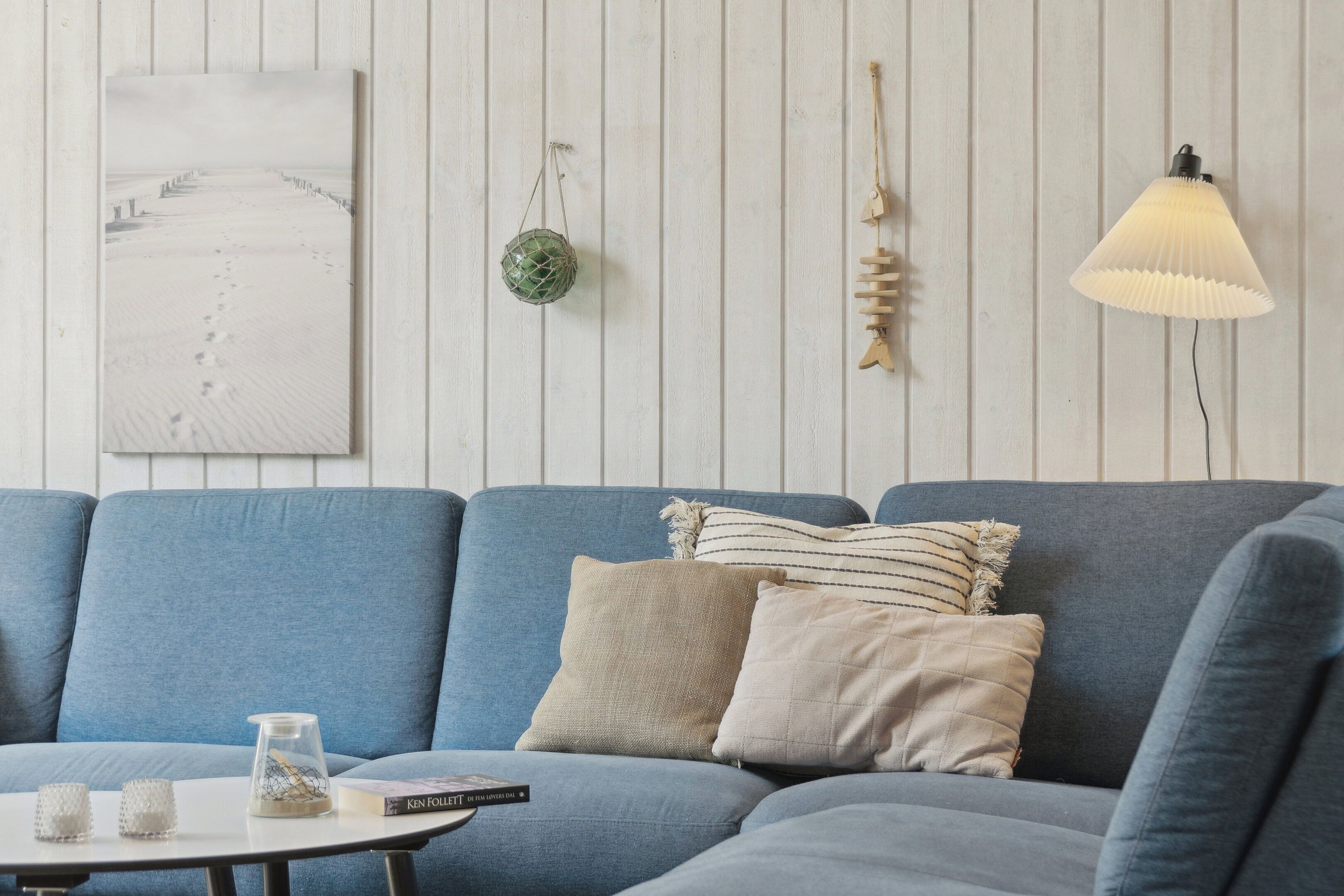 Et værelse med en blå sofa, et hvidt bord med en bog og et stearinlys, samt vægdekorationer bestående af et indrammet billede, en grøn pynt og en væglampe.