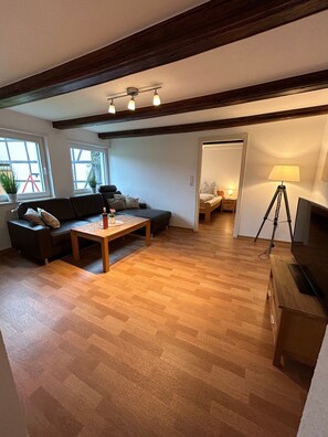 A spacious living area with a wooden floor, a sofa, a coffee table, and a TV stand. There are windows with plants and a door leading to another room.