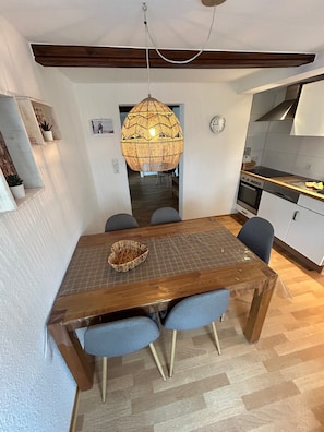 A dining area with a wooden table, blue chairs, a hanging light fixture, and a kitchen in the background.