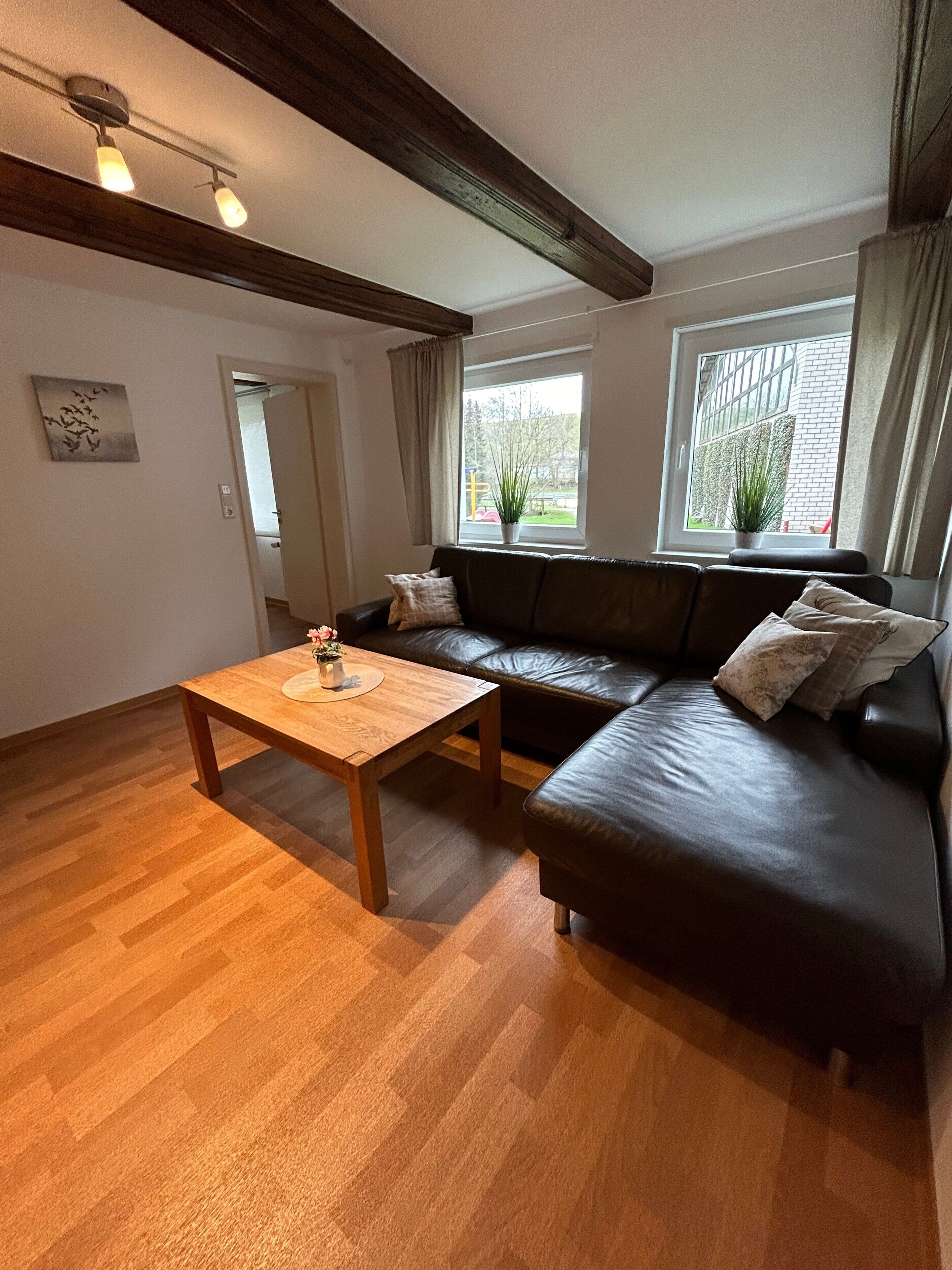 Een woonkamer met een houten vloer, een donker leren bank, een houten salontafel en een raam met uitzicht op groen.