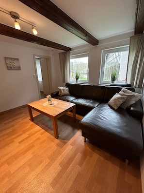 A living room with a wooden floor, a dark leather sofa, a wooden coffee table, and a window with a view of greenery.