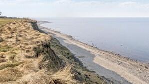 Un paysage côtier avec une falaise herbeuse, une plage de galets et une mer calme.