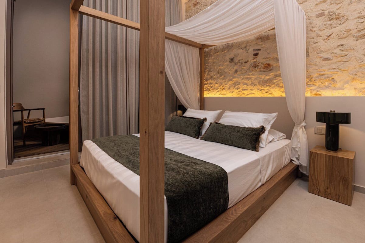 Una cama con dosel adornada con una tela colgante, una mesita de noche con lámpara y vista a un muro de piedra.
