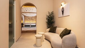 Habitación de hotel moderna con un sofá curvo, dos pequeños taburetes, una planta grande en maceta y una cama con vistas a un paisaje desértico.