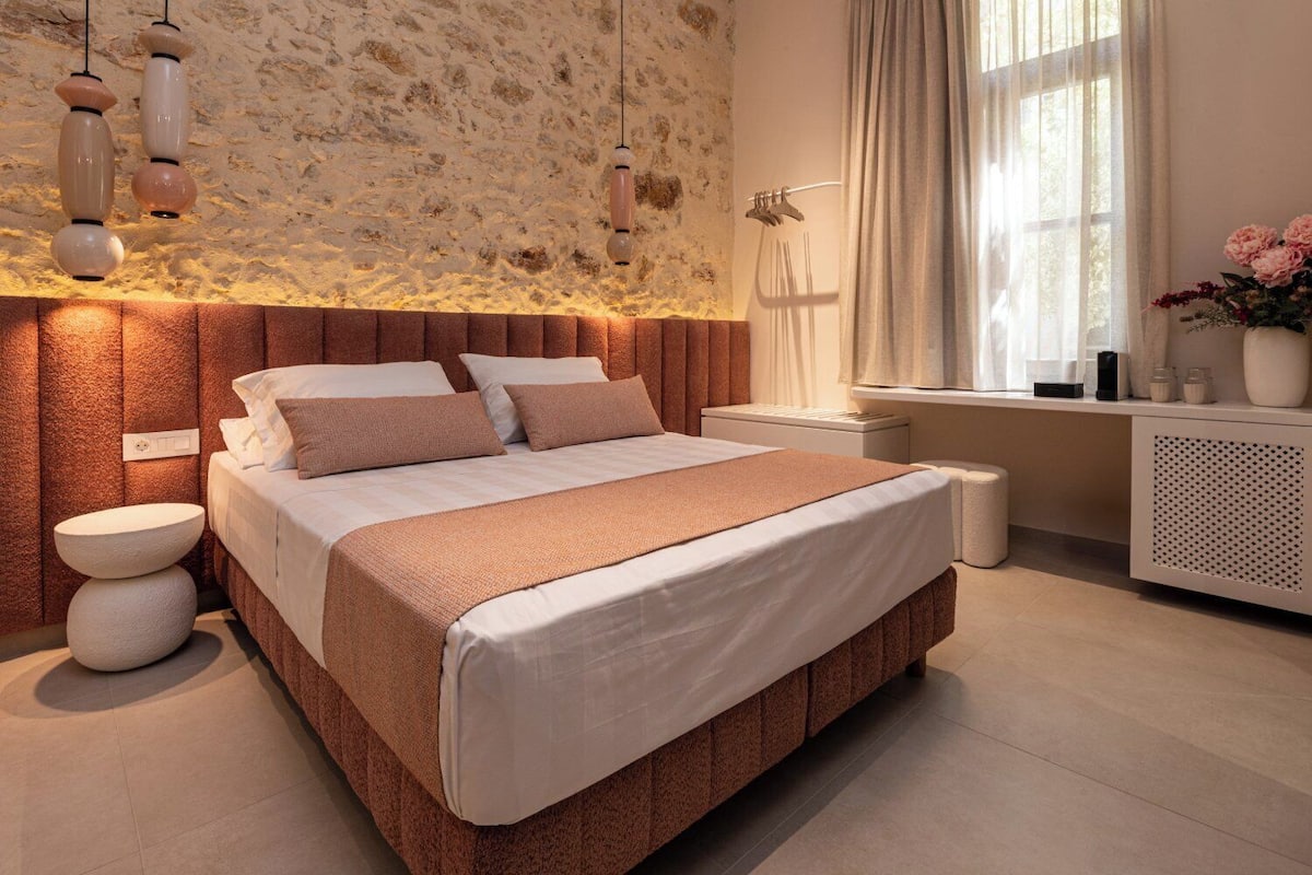 Un dormitorio moderno con pared de piedra, una cama grande con una decoración en tonos beige y marrón, mesitas de noche y una ventana con cortinas.