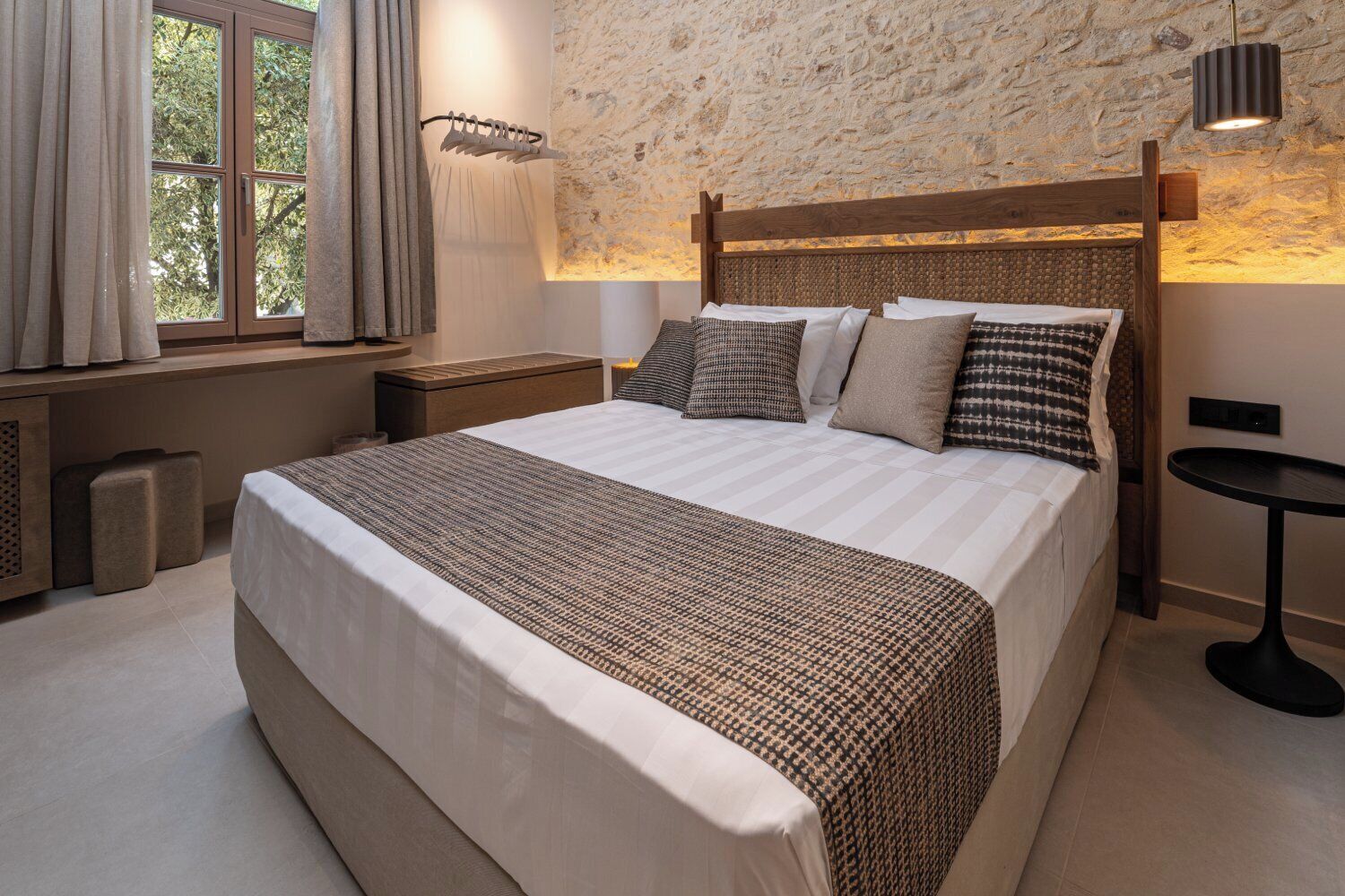 A bedroom with a large bed, a nightstand, a small table, and a stone wall.