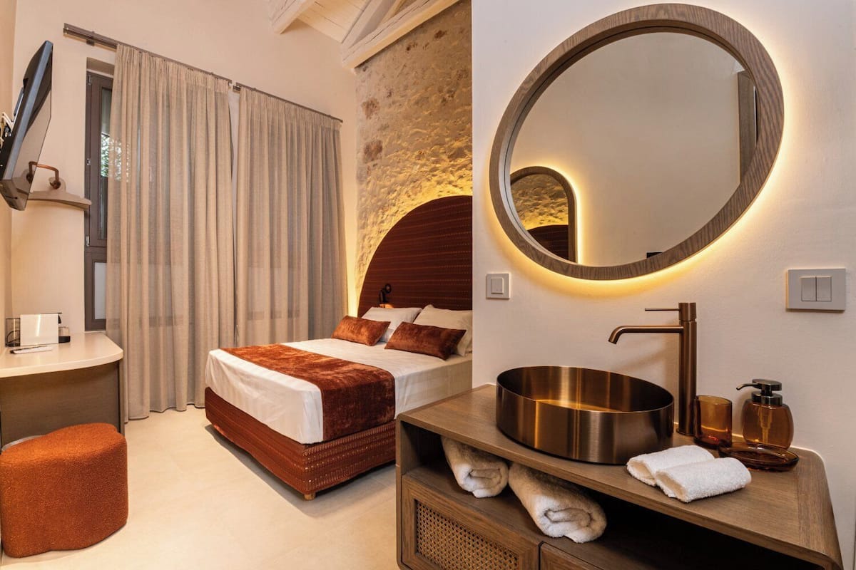 Una habitación de hotel moderna con una cama grande, un espejo redondo, un tocador de madera y una pared decorativa de piedra.