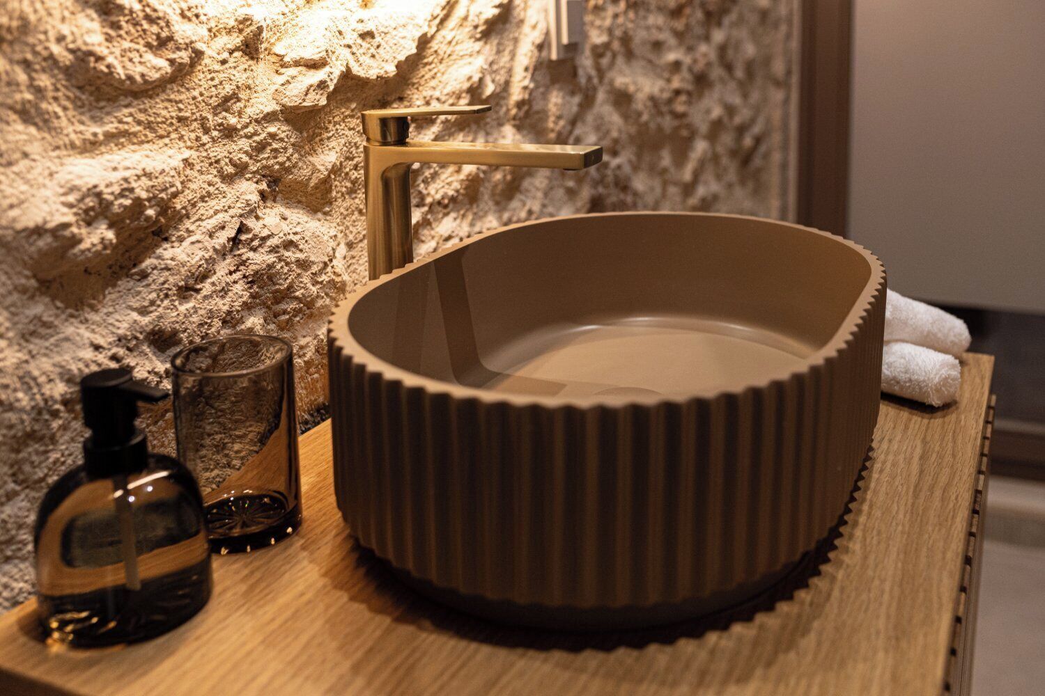A bathroom with a wooden vanity, a round sink with a ribbed design, a gold faucet, a glass bottle, and a glass.