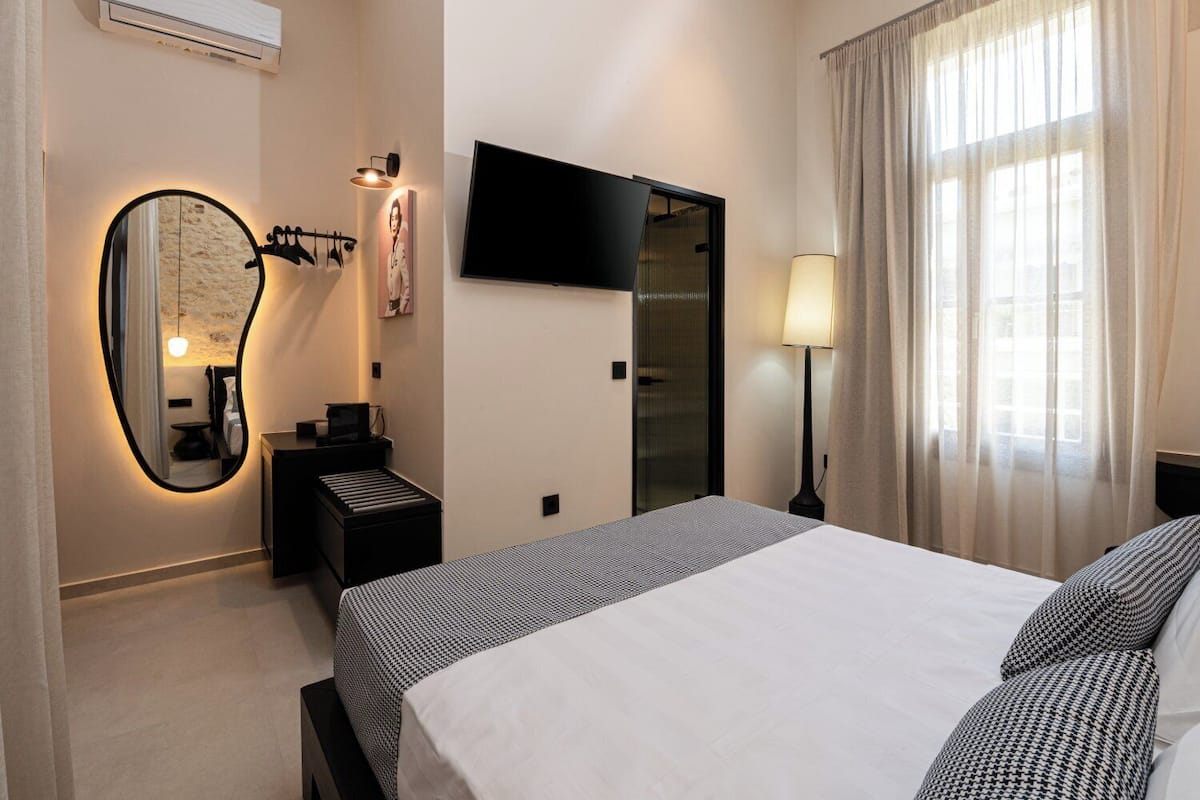 Una habitación de hotel con cama, escritorio, espejo, televisor y ventana con cortinas.