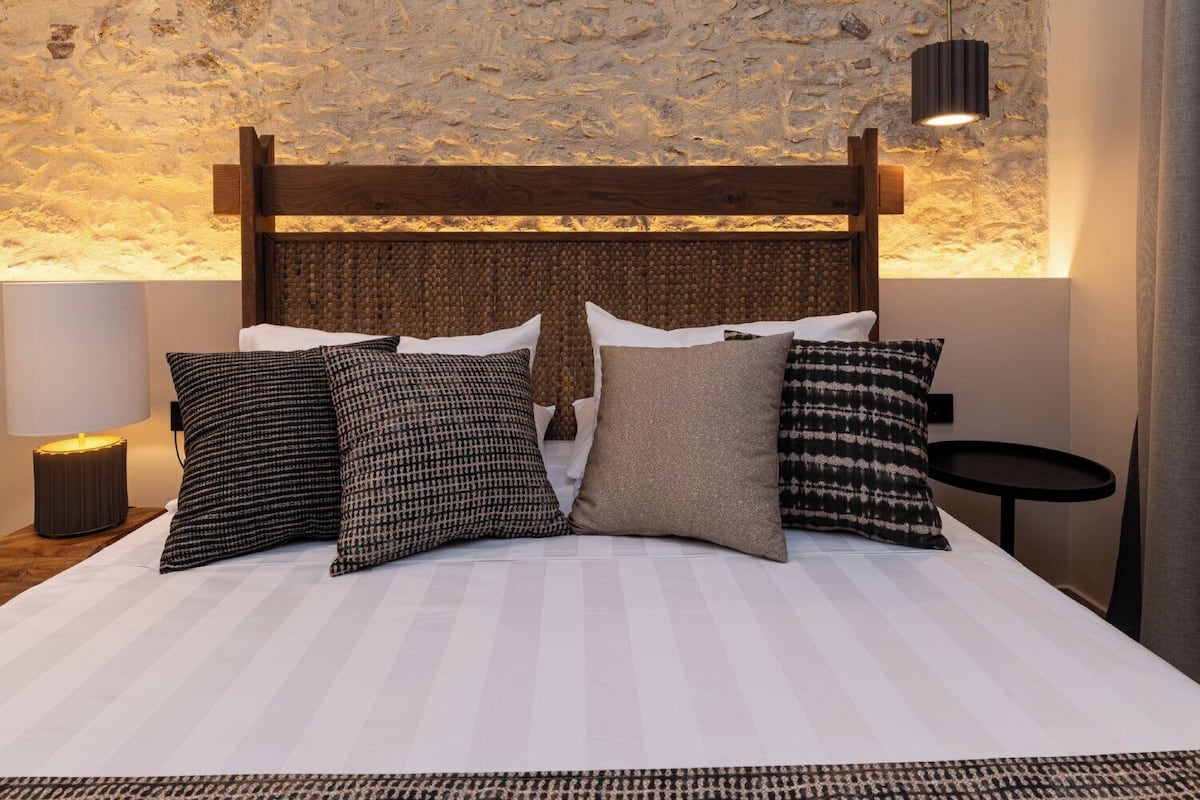 Una cama bien hecha con tablero de madera, varios almohadones, una lámpara de noche y una mesita redonda.