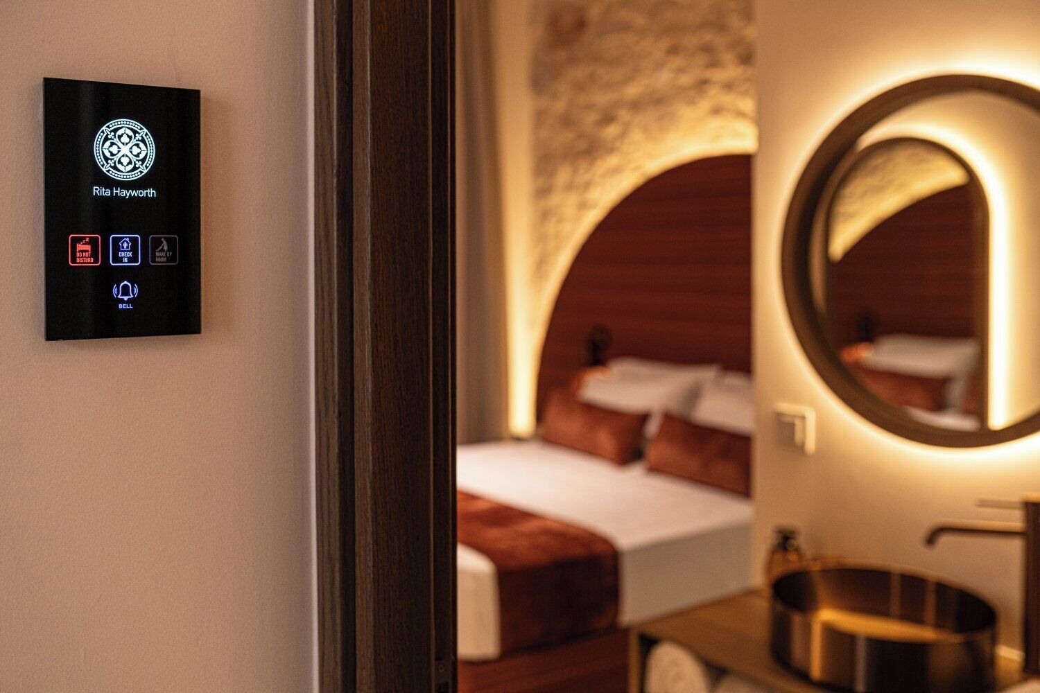A hotel room with a bed, a round mirror, and a wall-mounted control panel with a logo and buttons.