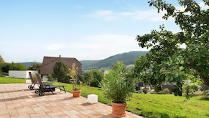 Un patio con mobiliario exterior, plantas en macetas y vista a una casa y colinas al fondo.