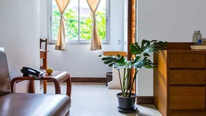 Quarto com cadeira de madeira, escrivaninha com telefone, planta em vaso, janela com cortinas e armário de madeira com garrafas.