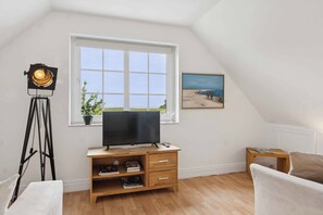 Ein Zimmer mit Fernseher auf einem Holzständer, einem Stativ, einem Fenster mit Meerblick und einem Gemälde an der Wand.