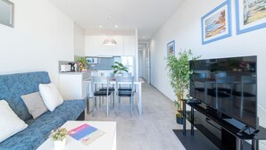 A modern living room with a blue sofa, a dining area with chairs and a table, a kitchen in the background, and a TV on a stand. - Apartment in Empuriabrava with Sea Views (Empuriabrava)