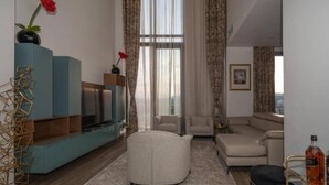 A modern living room with a sofa, a coffee table, and a TV on a blue cabinet. There is a window with curtains and a painting on the wall. - Africa's Touch & Leisure (Kigali)