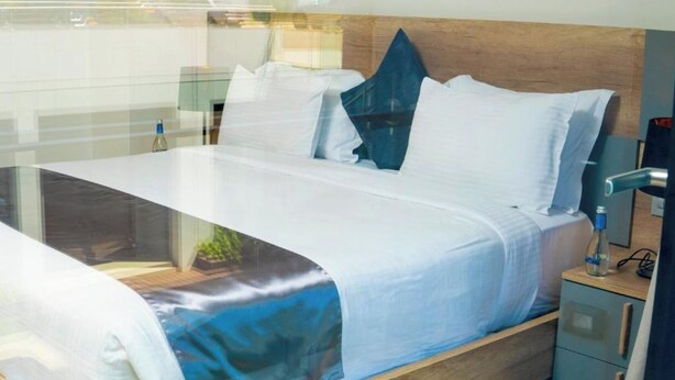 A modern hotel room with a wooden headboard, a glass partition, and a bed with white bedding. - Africa's Touch & Leisure (Kigali)