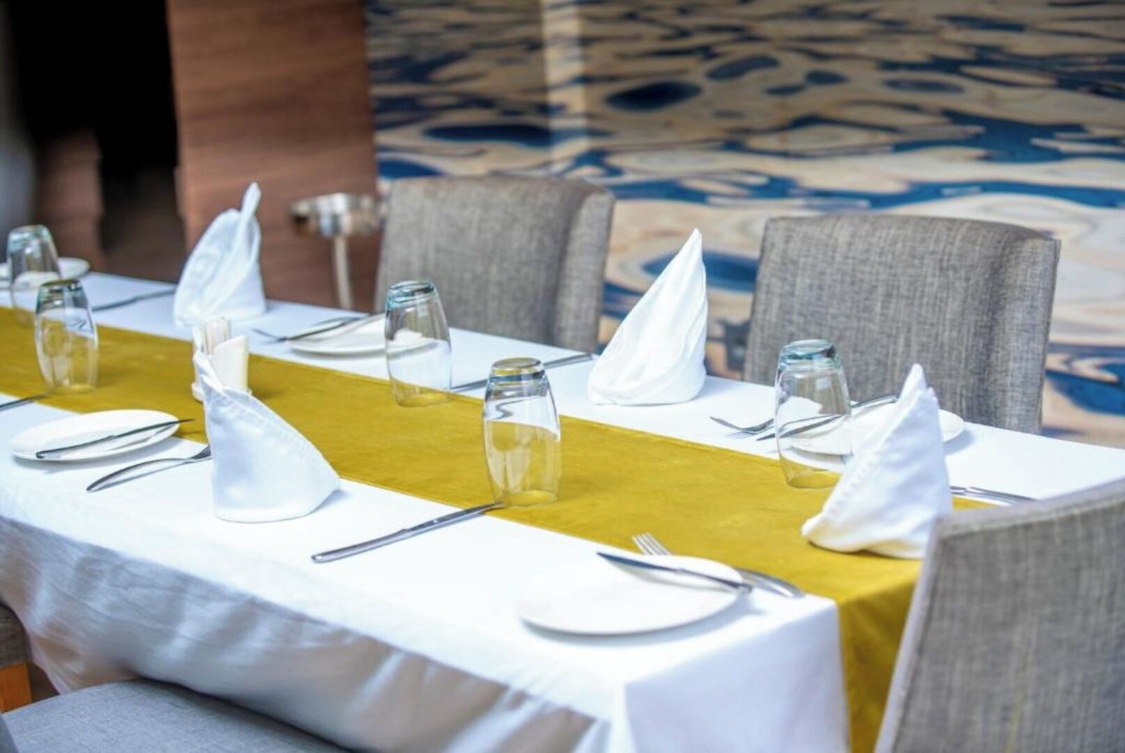 A neatly set dining table with white tablecloth, yellow runner, and grey chairs.