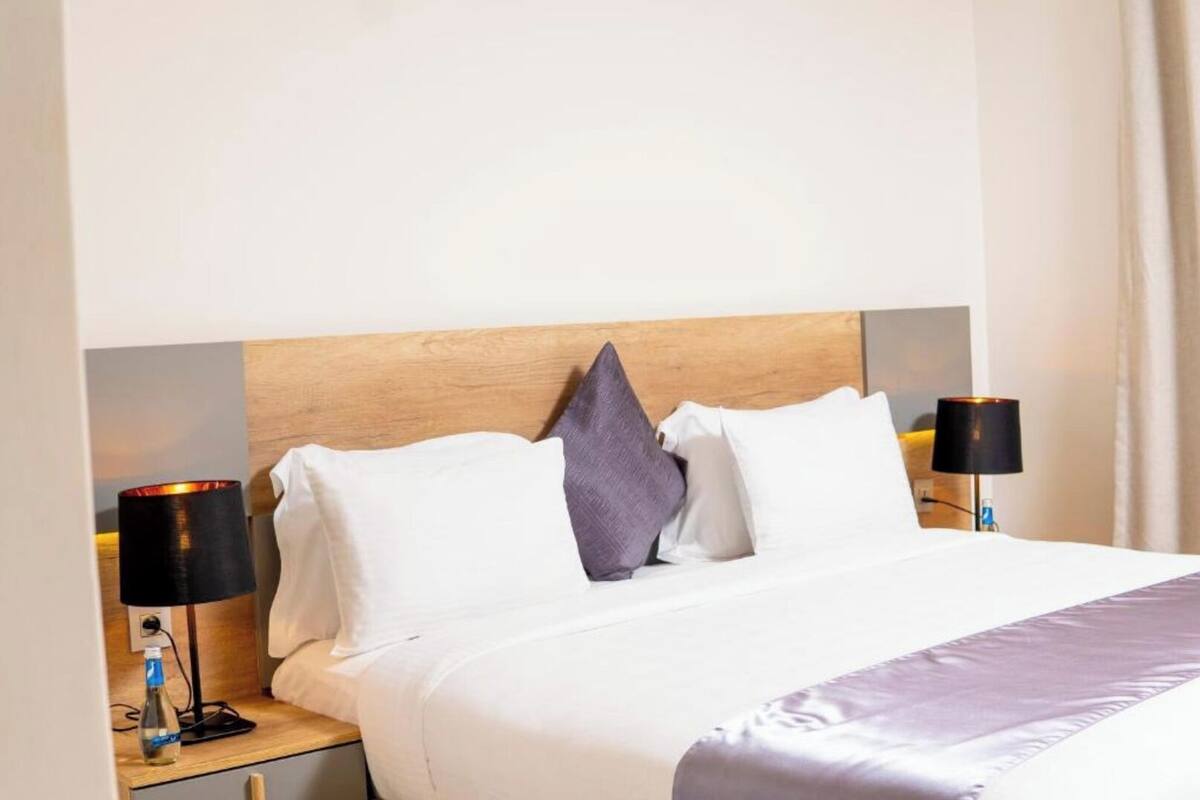 A neatly made bed with white linens and a wooden headboard, flanked by two bedside tables with lamps and a bottle.